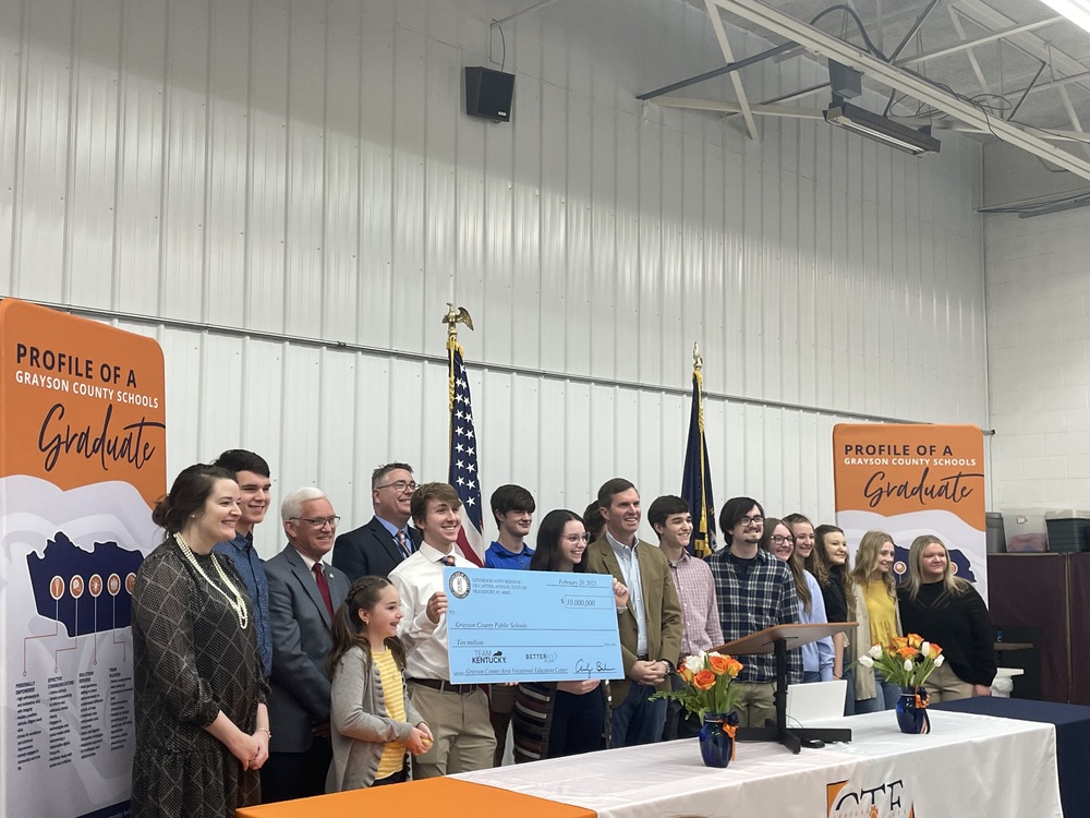 GCS students, Supt Doug Robinson with Gov. Beshear , Steve Meredith and Samara Heavrin holding a large ceremonial check standing in front of an orange, white and navy draped table with CTE logo. American and KY flags behind and flanked by Profile of a GCS graduate vertical banners.