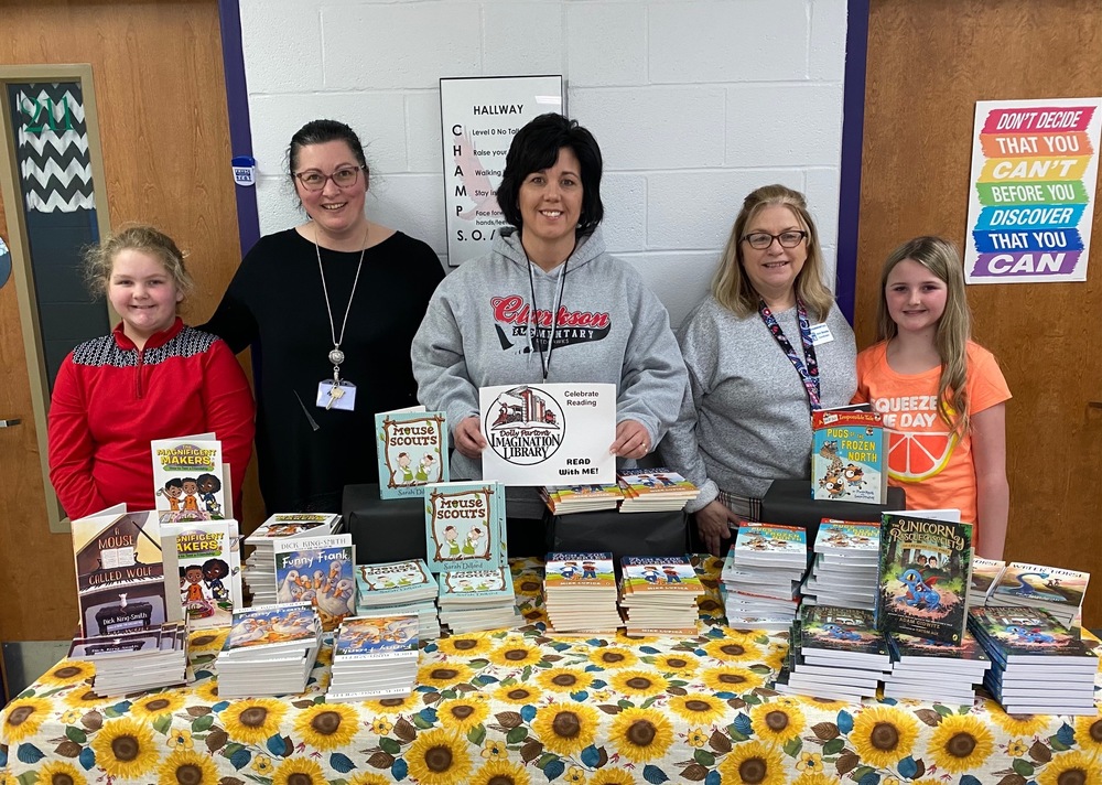 2 students flank FRC Director Ann Brooks, assistant FRC director and library media specialist Kim Keys in front of a sunflower tablecloth draped table with stacks and upright books on it