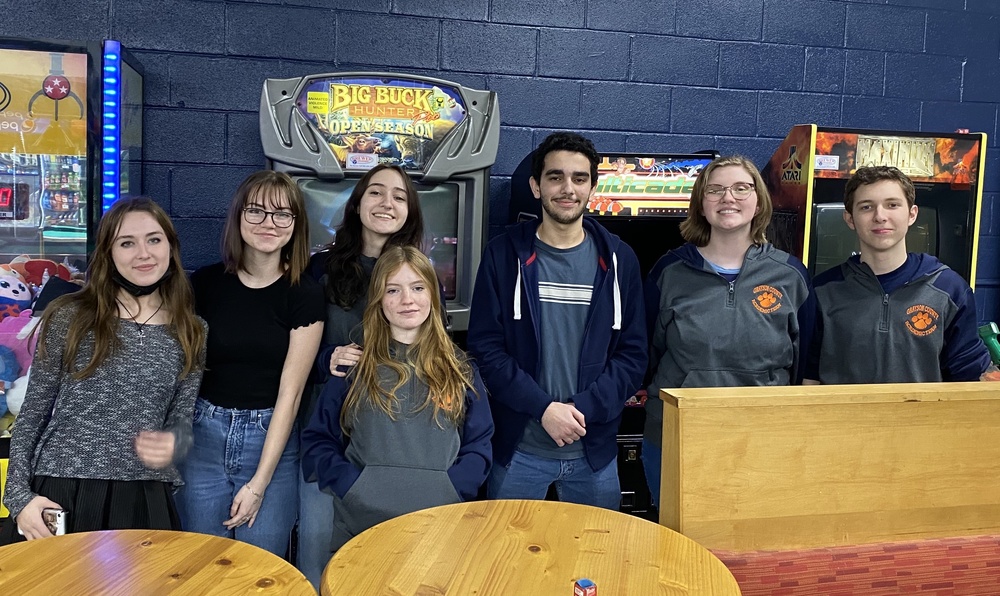 GCHS Academic Team members standing in a row in front of arcade machines, behind round wooden tables and a tall booth bench