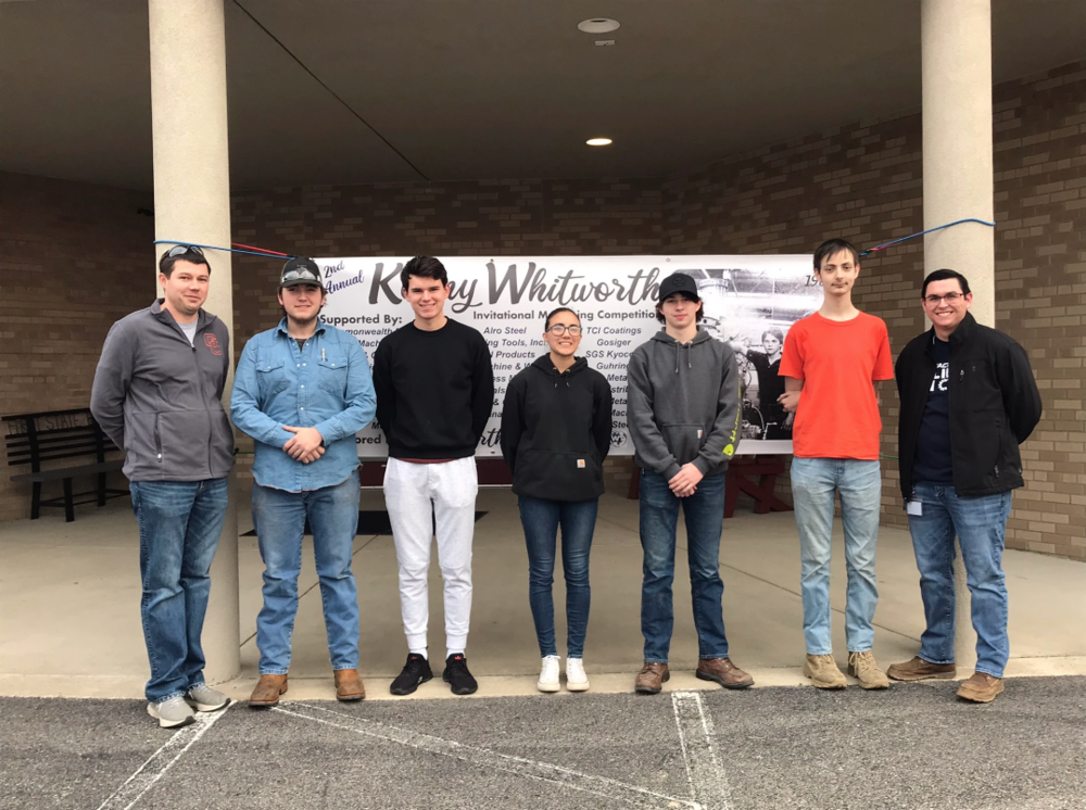 Outside photo - Machining teachers Michael Critchelow (left) and Josh Walters (right) flank  5 GCHS machining students (two male on either side of female) in front of Kenny Whitworth Invitational Banner between two columns