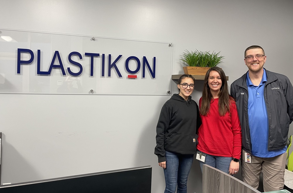 Man in blue shirt and grey jacket standing to the right of dark-haired woman in red pullover and shorter woman in black pullover. Plastikon logo on clear acrylic sign to their left