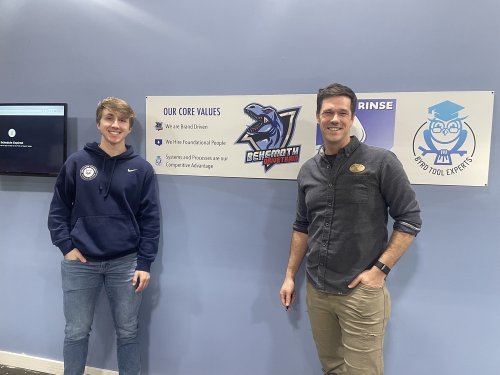 Young man in navy hoodie and jeans standing to left of man in khakis and grey pullover in front of blue wall with Behemoth company logo and core values banner behind them. 