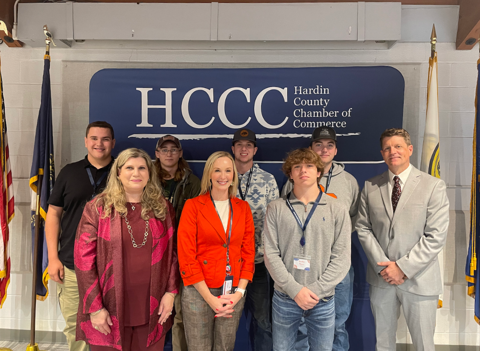 CTE students and sponsors (three women, one male in front, five males in back) in front of Hardin County Chamber of Commerce Sign - white letters on dark blue background. American and KY flags to left and white and navy flag (unidentified) to right