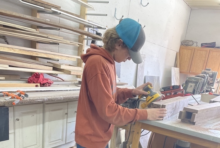 Wyatt Clemons working on a carpentry project at a table with wooden slats, more behind him on shelves.