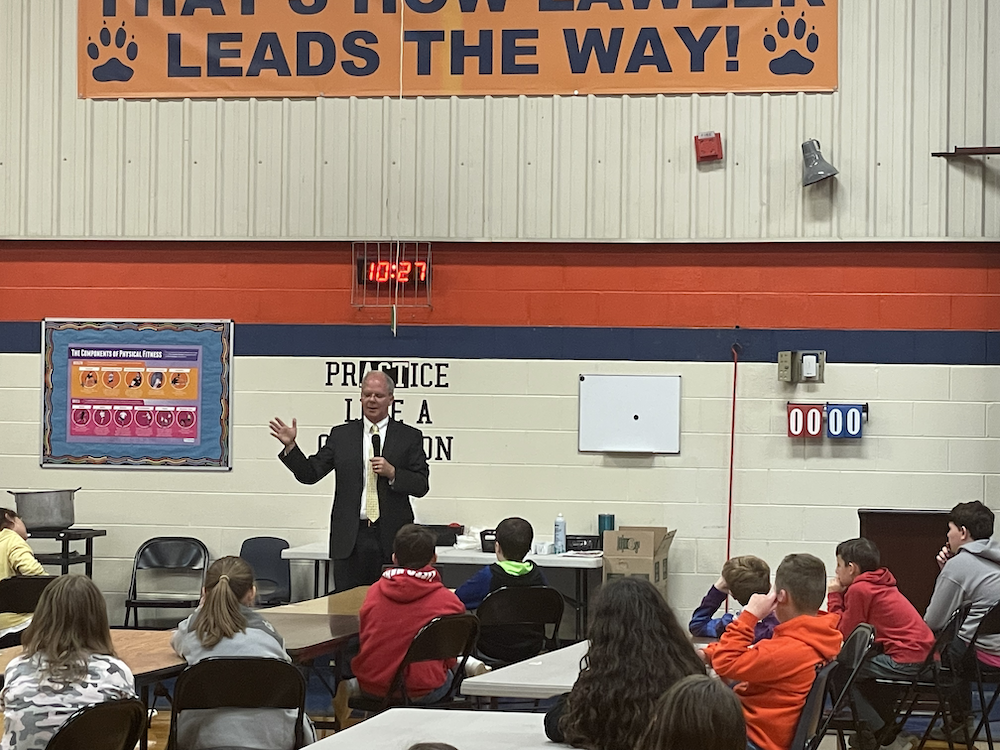 Rep Brett Guthrie talking to students in the Lawler gym with an orange "leads the way" banner and PE bulletin board behind him. They are seated by desks and a