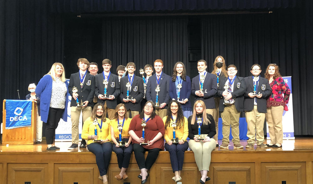 DECA members on stage at Regionals, most in DECA blazers, holding trophies, one row sitting, one row standing behind