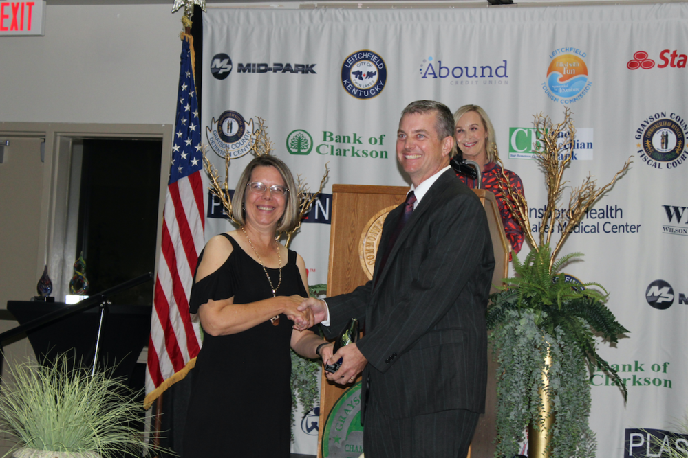 Matt Hayes receiving an award from Chamber president Jennifer Priddy as Brandi Lee looks on from podium in front of Chamber Dinner Sponsors step and repeat banner