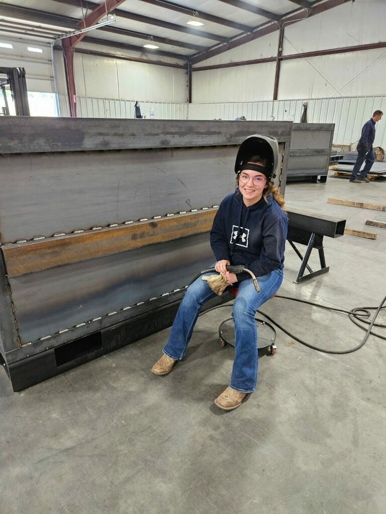 Addison Newton seated near a metal wall in welding helmet
