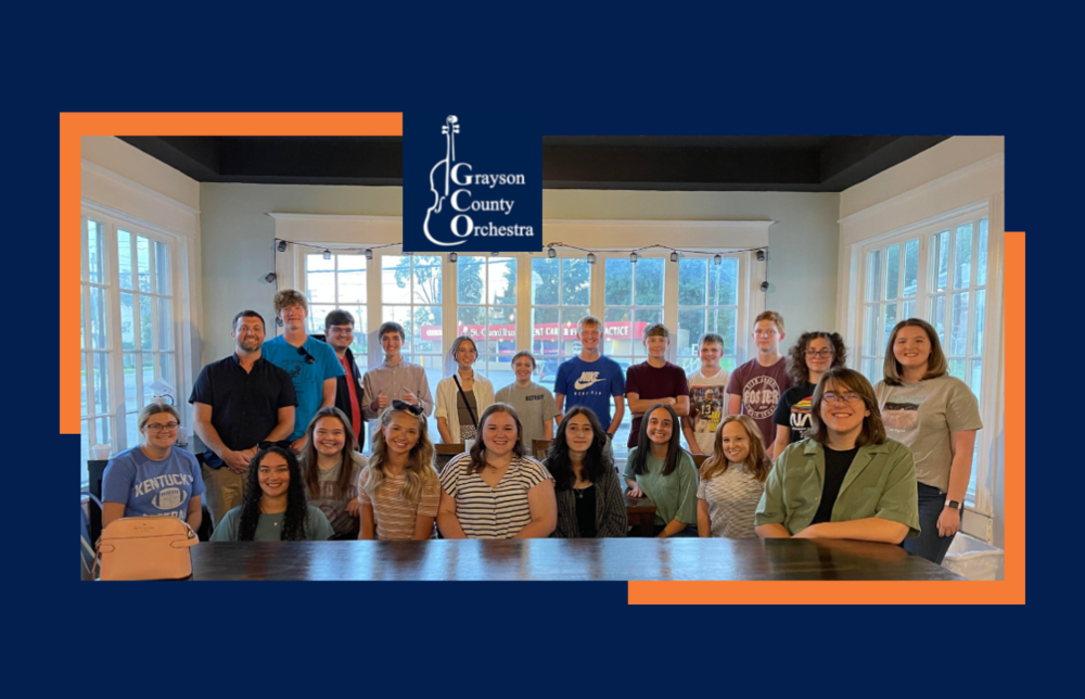 GCHS Chamber Orchestra, Directors Adam and Stacy French witting at a table in front of windows surrounding on all sides. GC Orchestra logo image featured at top center es