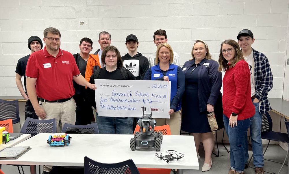 TVA, WRECC reps, GC Tech Center students and staff holding large ceremonial grant check standing in front of table with a lego and a tank robots and a small drone on it.