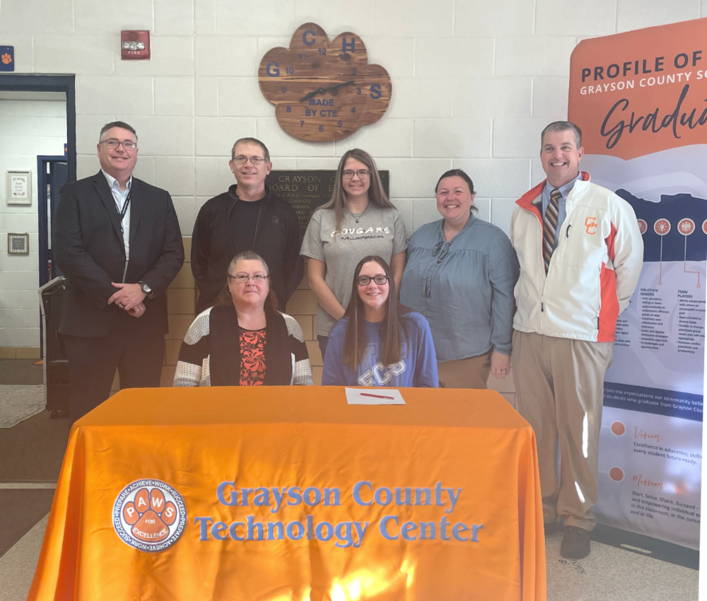 GC Supt. Doug Robinson, Father and sister of Allie Dotson, FCS teacher Cody Mooneyhan, GC Tech Center Director Matt Hayes behind, Allie Dotson and mother in front of a GCTC orange cloth draped table with GCS Profile of a Graduate vertical banner to right side.