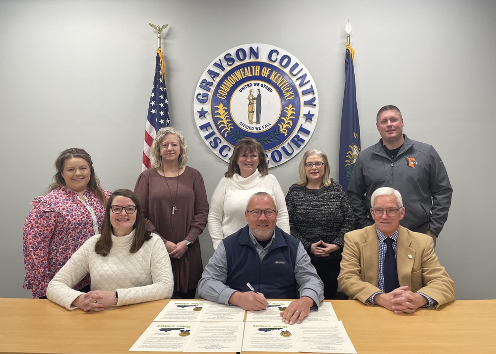 State Rep. Samara Heavrin, Judge-Executive Kevin Henderson with proclamations before signing, and State Sen. Steve Meredith all seated at a table. Back standing: Shalee Watson, Kim Cannon, Barbara Allen, Ann Brooks, and Josh Baldwin, all with GC Schools. American flag, GC Fiscal Court logo and KY flag behind