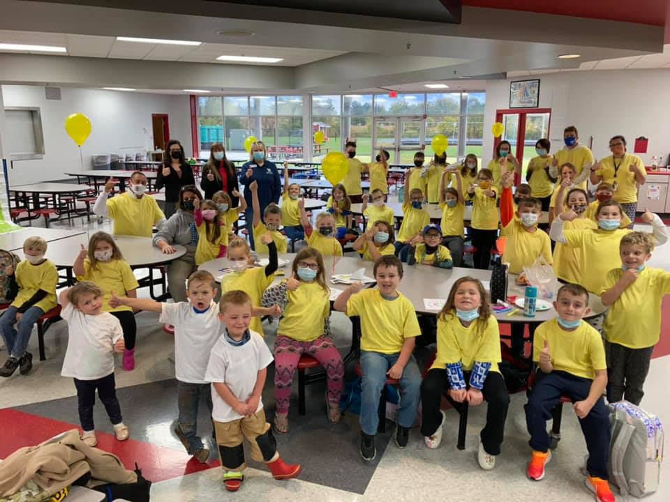 children and adults in mostly yellow and a few white thsirts in a lunchroom with hands up 