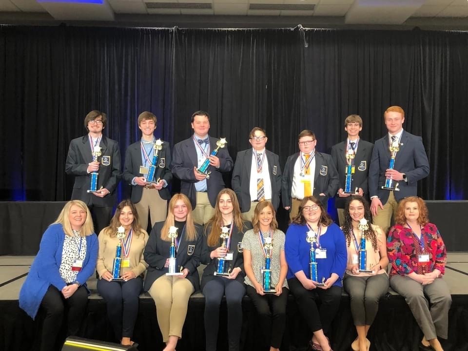 GCHS DECA members and sponsors on stage with trophies, one row sitting, back row standing, all in DECA blazers