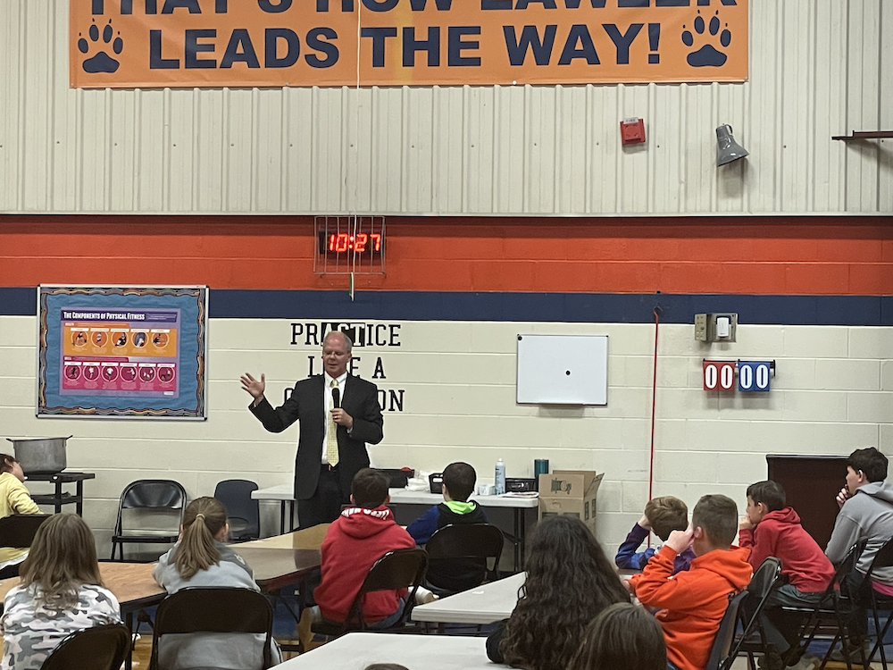 Rep Brett Guthrie talking to students in the Lawler  gym with an orange "leads the way" banner and PE bulletin board behind him. They are seated by desks and a 