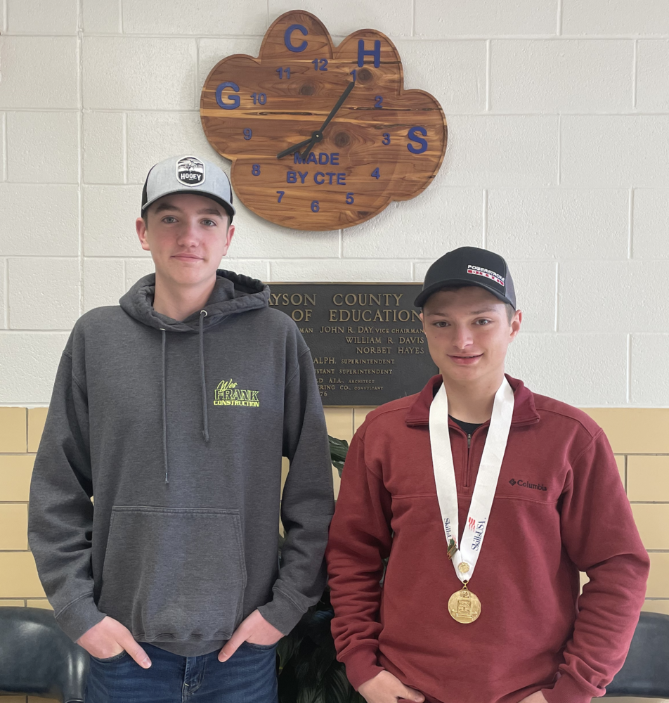 Paul Overton and Thane Smith standing in from of GCHS wooden cougar paw clock. Smith, on right has award medal on white ribbon around his neck
