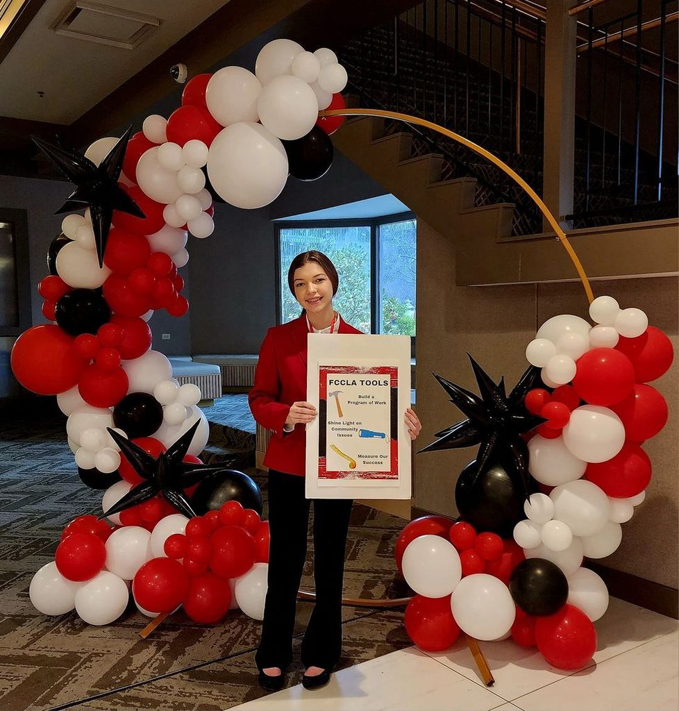 Michenna Meredith in a red FCCLA blazer and black pants holding a red and black poster in front of a red, white and black balloon sculpture 