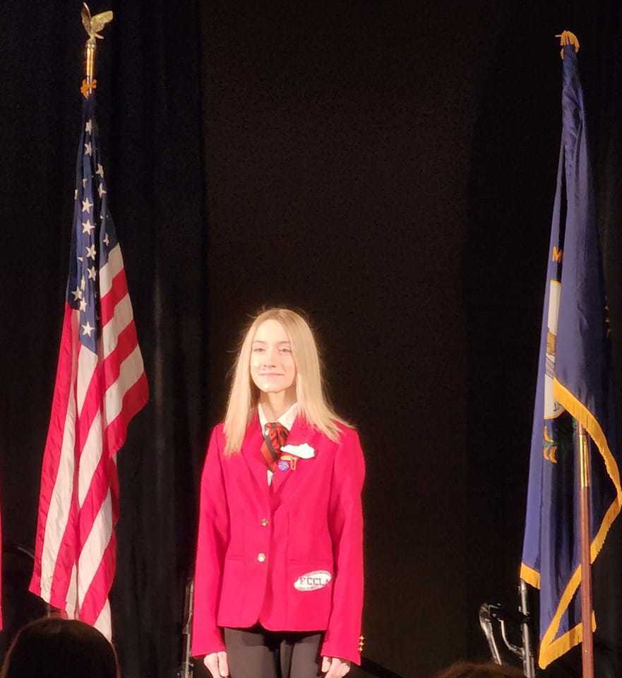 Hollie Hagan on stage in red FCCLA blazer between  American and KY Flags